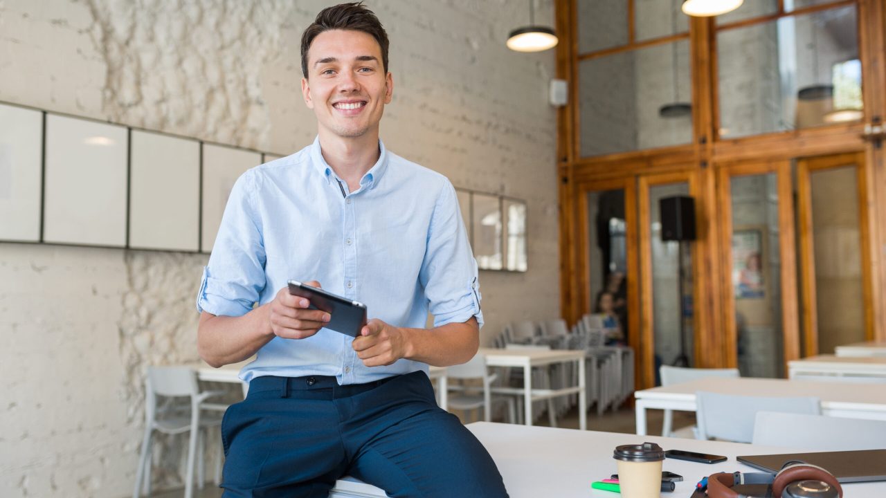 young stylish smiling man in co-working office, startup freelancer holding using tablet, modern office room workplace, student education online